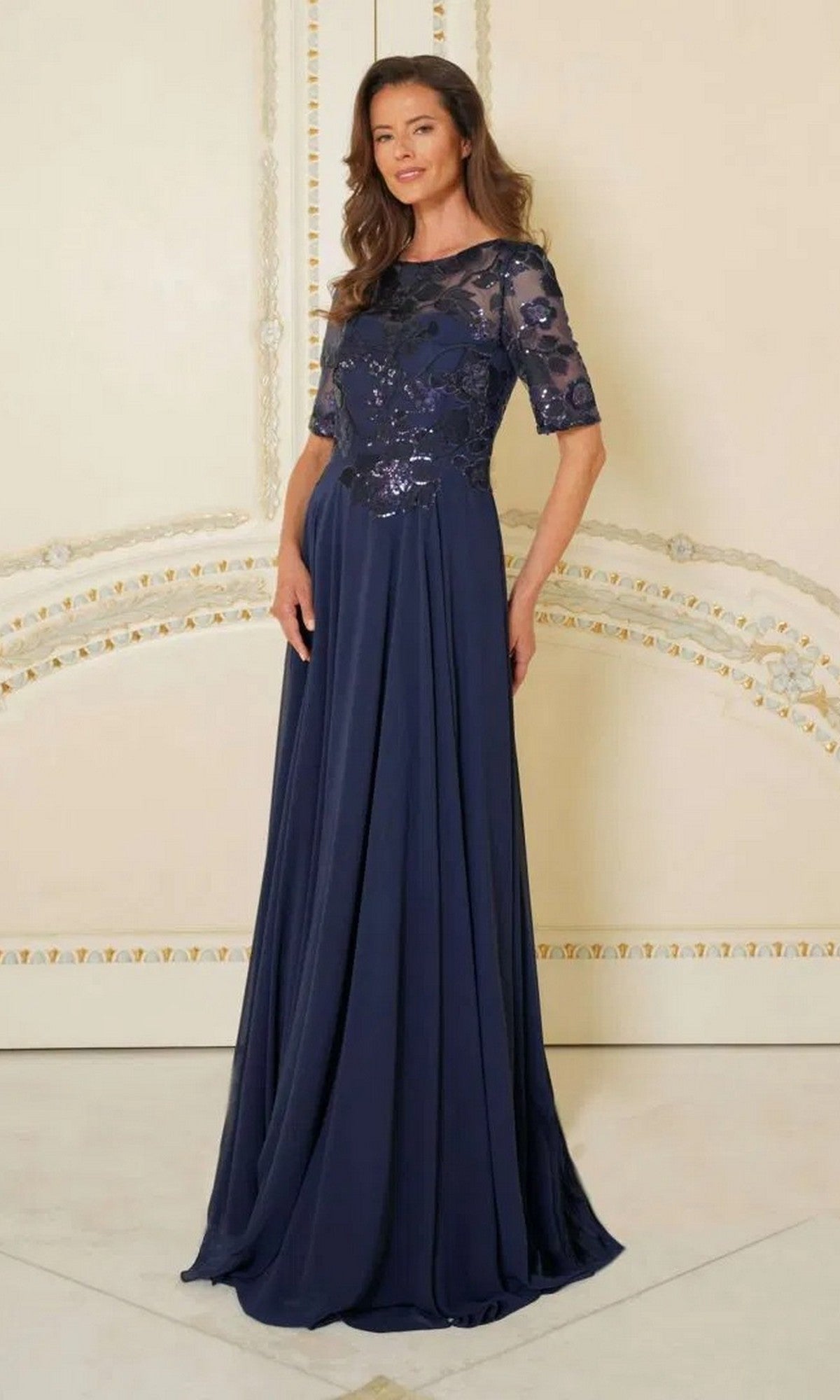 A woman stands before an ornate light-colored wall wearing the Marsoni M286, a long navy formal dress featuring a sheer sequin lace bodice and short sleeves.